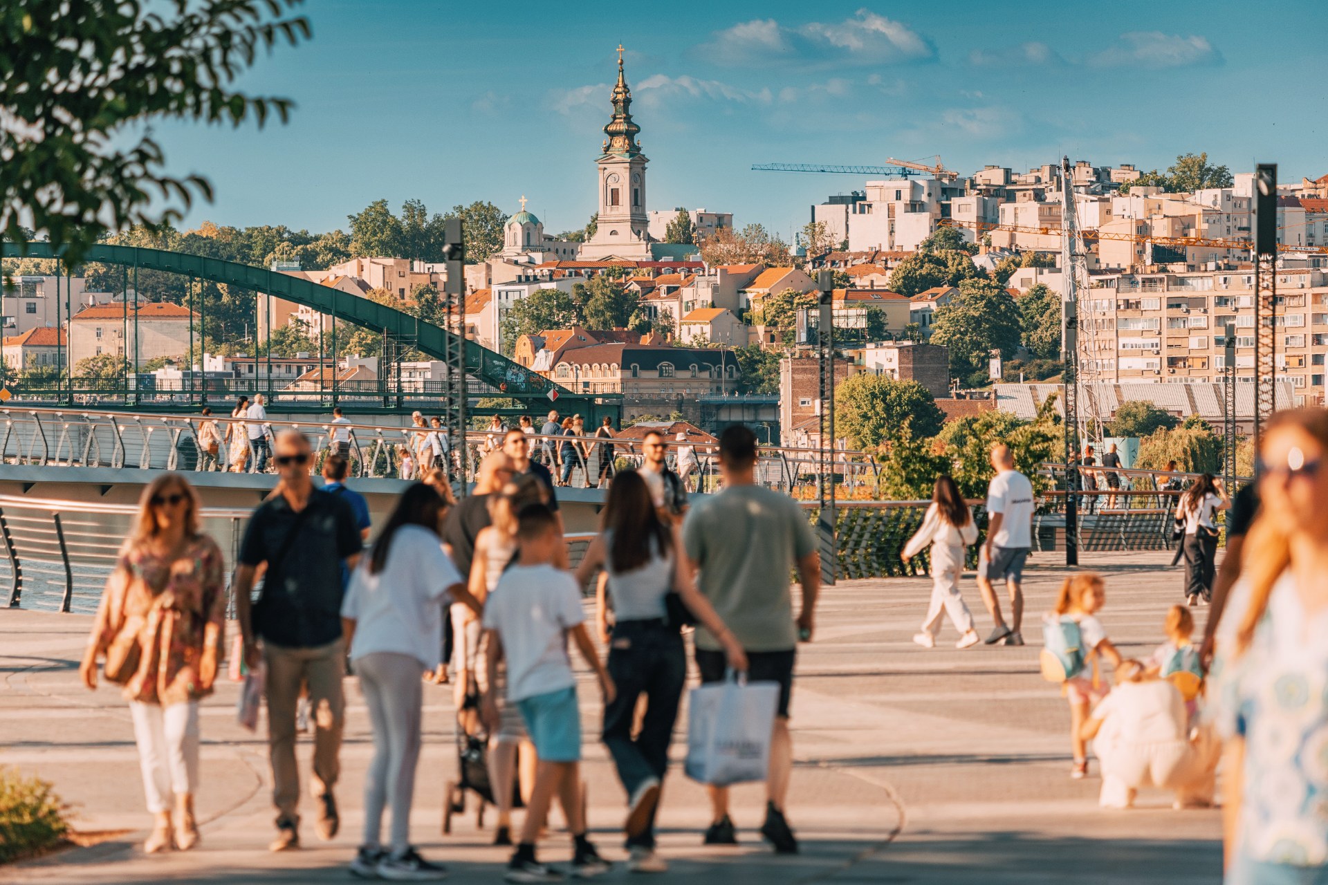 Menschen spazieren auf der Save-Promenade in Belgrad, dahinter die Altstadt und ein Kirchturm